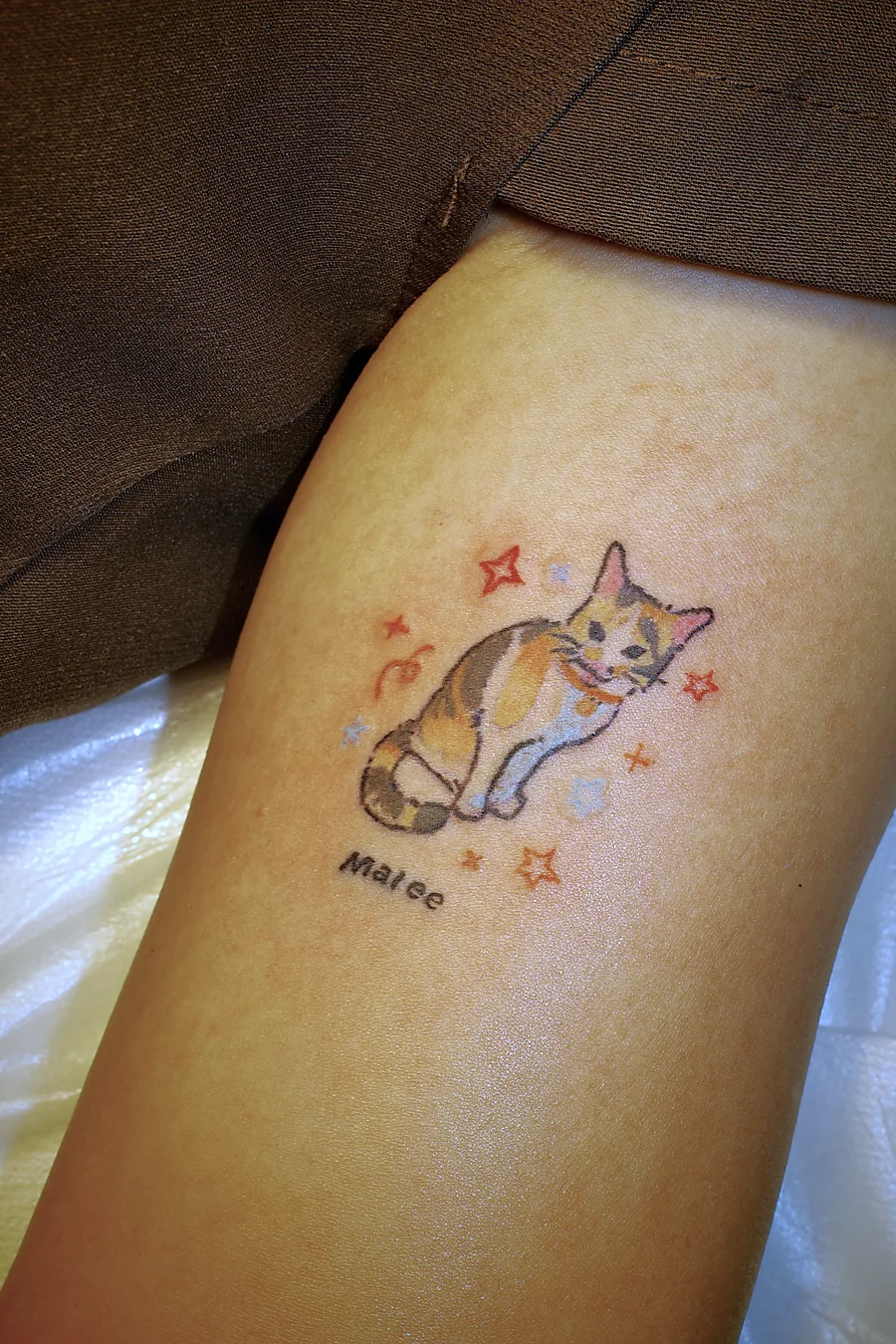 Calico cat portrait tattoo named Malee on the thigh, a colourful naive-style cat licking its paw surrounded by red, orange and pale blue stars