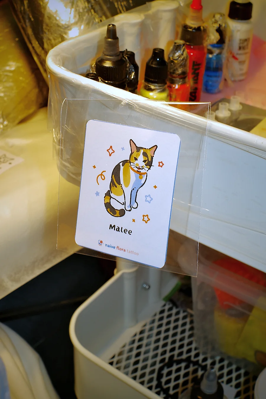 The Malee cat design printed as a Naive Flora card, clipped next to the ink bottles at the tattoo station