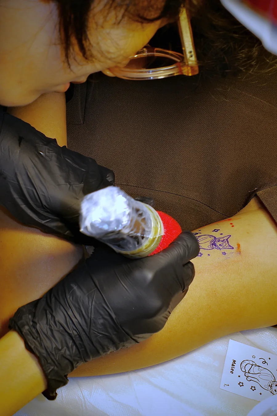 Close-up of gloved hands tattooing the calico cat outline on the client's leg