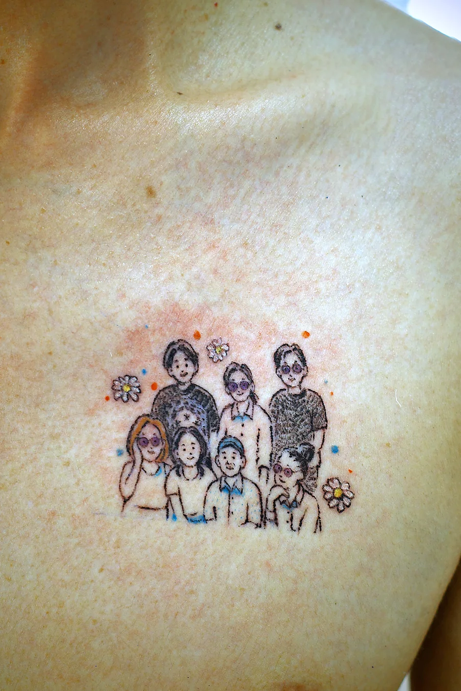 Family portrait tattoo on the upper chest: seven small naive-style figures with sunglasses, a cap and daisies, fresh from the session